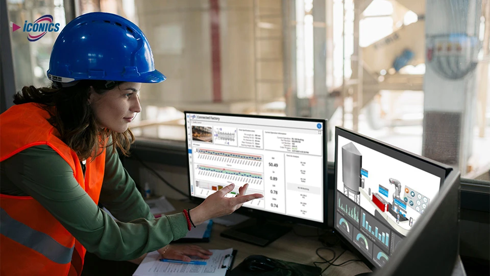 Photo of woman with a hard hat and orange safety vest leaning over a desk with three monitors featuring dashboard and schematic views
