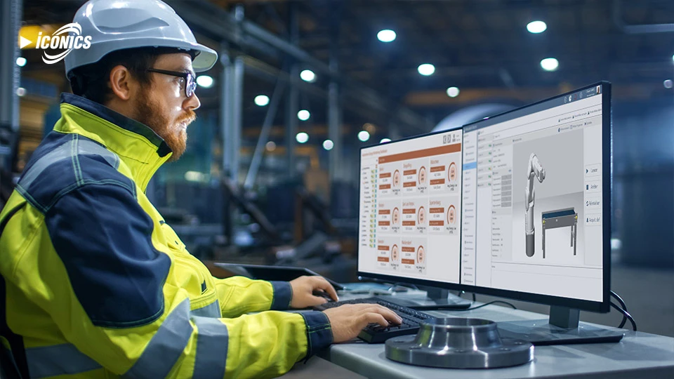 Photo of man wearing a hard hat and high-vis safety jacket sitting at a desktop with two monitors showing dashboards and a rendering of the Mitsubishi Electric Robotic Arm