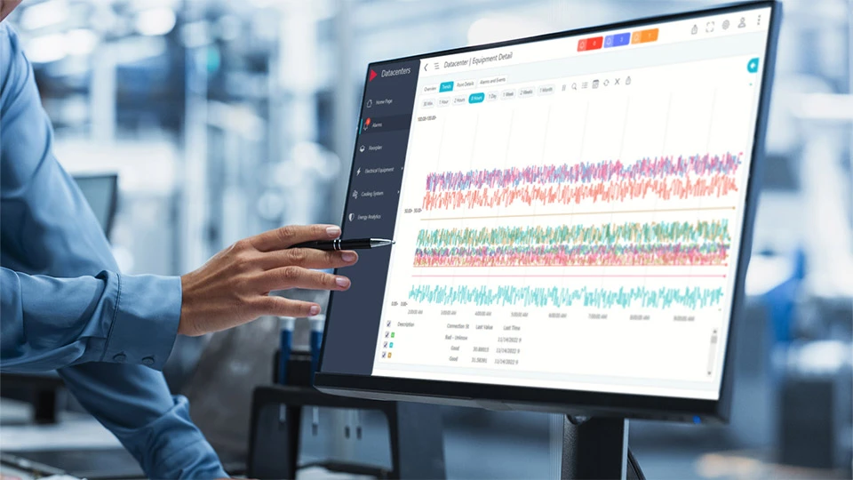 A person is pointing to a computer monitor displaying colorful, time-series data graphs. The screen shows a dashboard labeled “Datacenter / Equipment Detail,” with data trends and performance metrics 