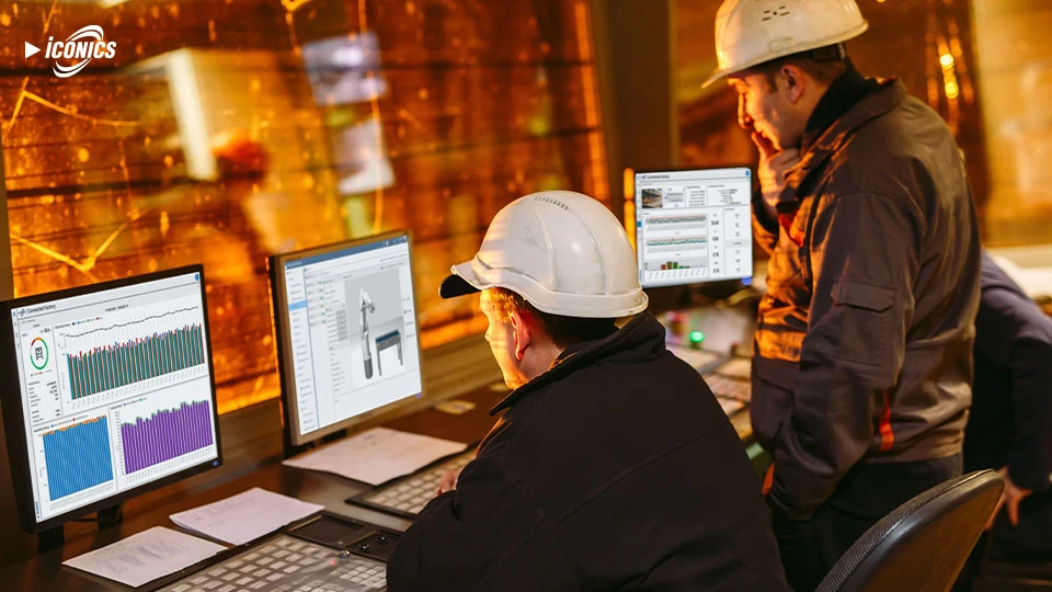 Photo of two men, one seated and one standing, in a control room facing monitors showing various dashboard views and one o the Mitsubishi Electric robotic arm