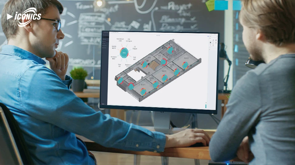 Photo of two men sitting at a desk in a classroom, in front of a monitor featuring a floor plan schematic, with a chalkboard in the distance