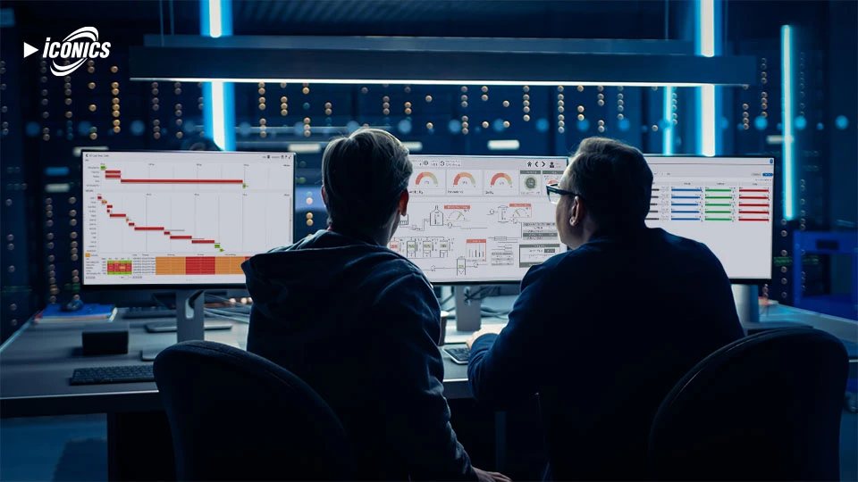 Two men in shadow sitting at a desk in a server room, with three monitors featuring various dashboard views